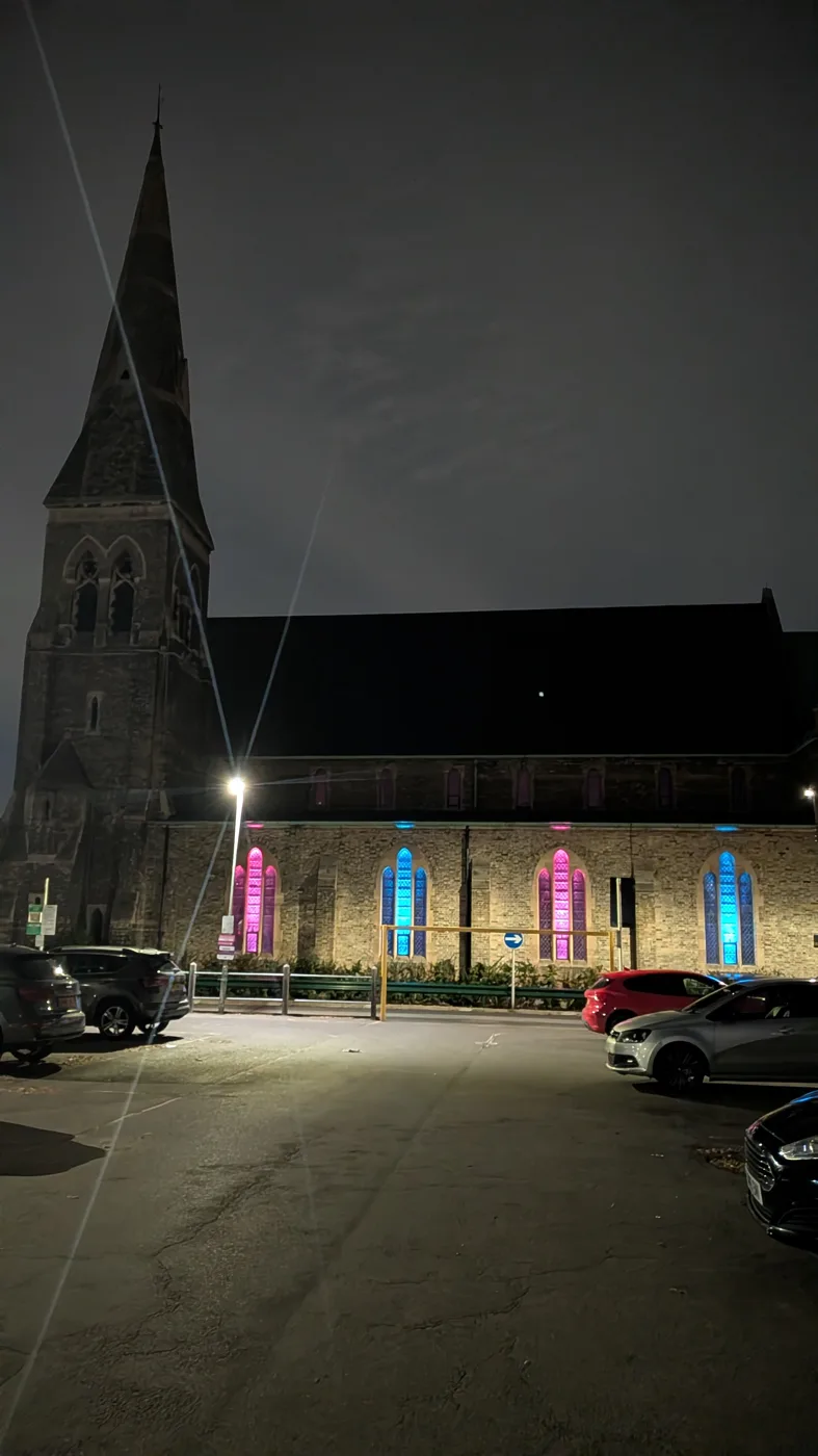 Christ Church Beckenham at night, its tall arched windows lit alternately pink and blue, with cars parked in front