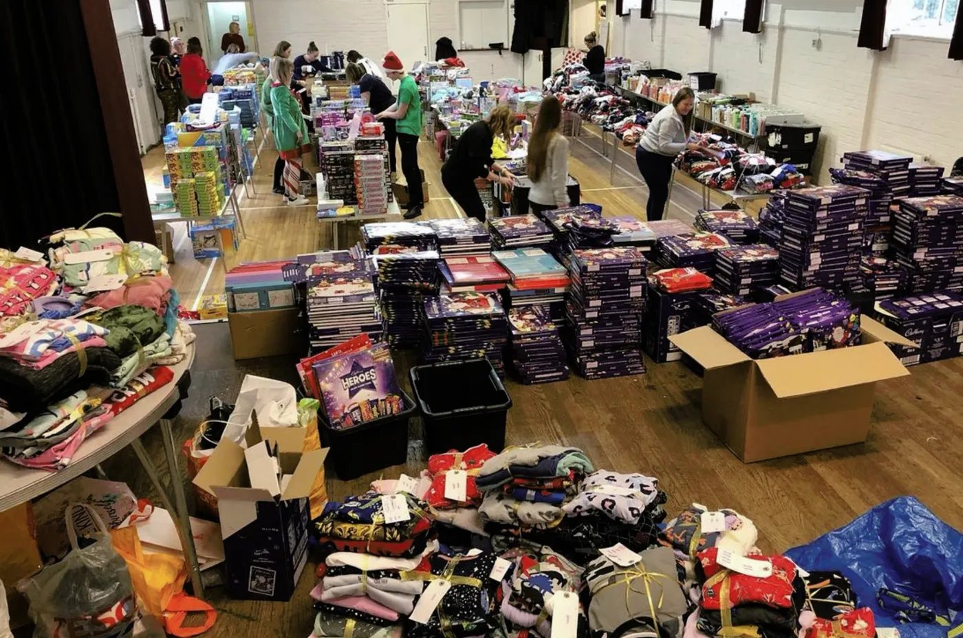 Volunteers working along tables in a church hall stacked with advent calendars, selection boxes, pyjamas and wrapped gifts, during a Bromley Brighter Beginnings packing day