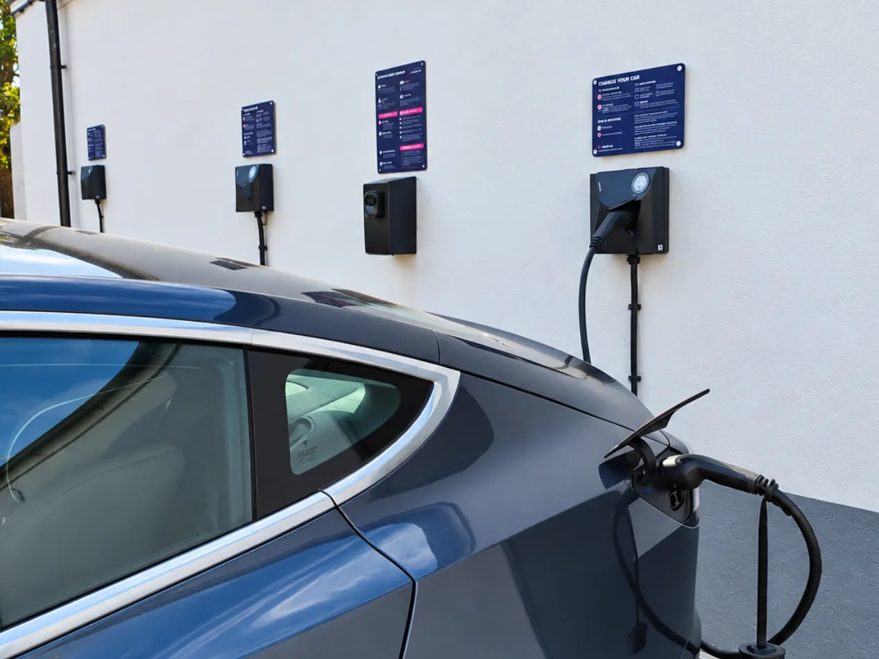 A grey electric car plugged into one of the wall-mounted chargers at The Bromley Court Hotel, with further chargers and instruction signs along the white wall behind