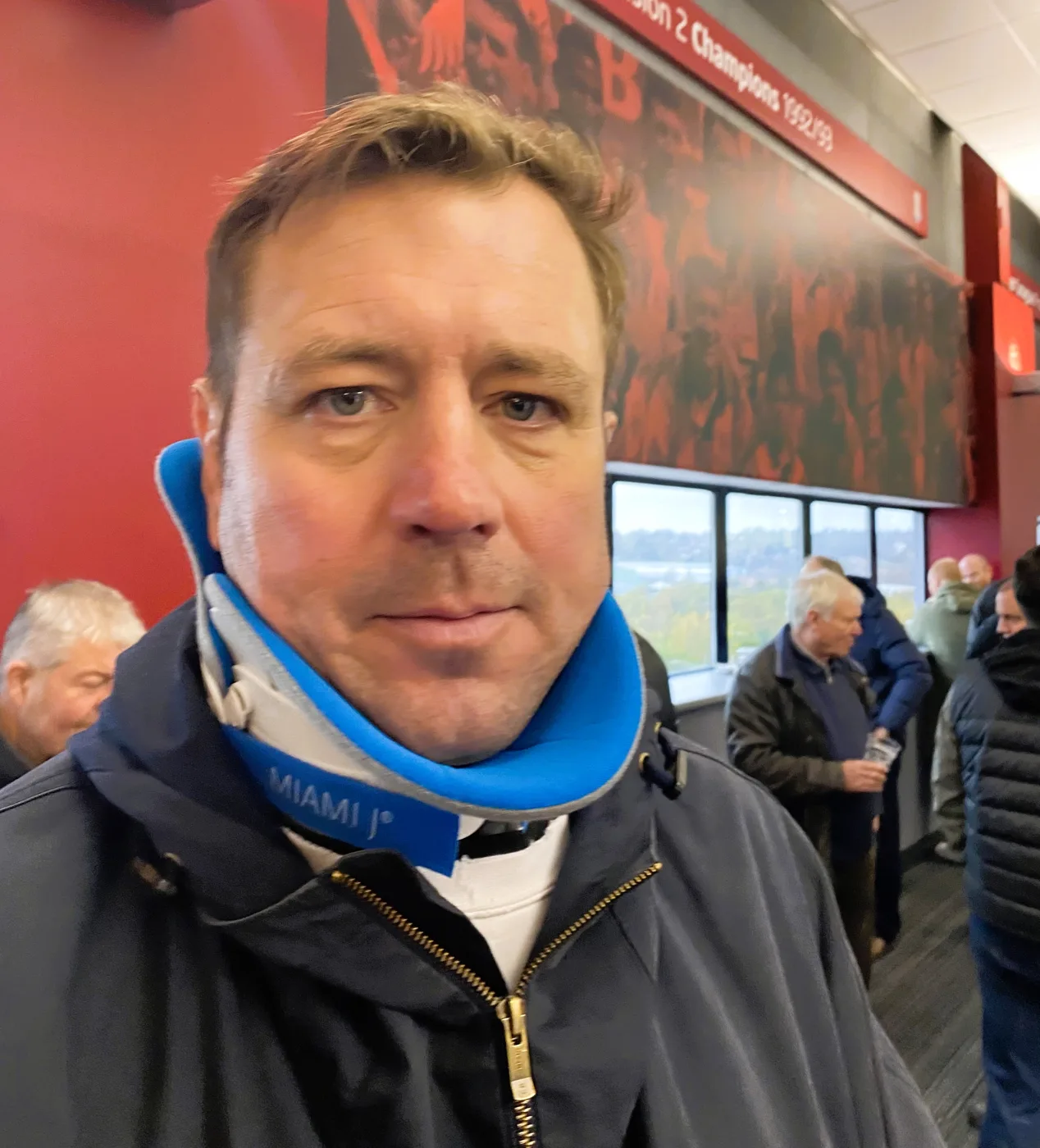 Peter Evans wearing a blue and grey Miami J neck collar under a dark jacket, standing in a red-walled stadium concourse with other people behind him