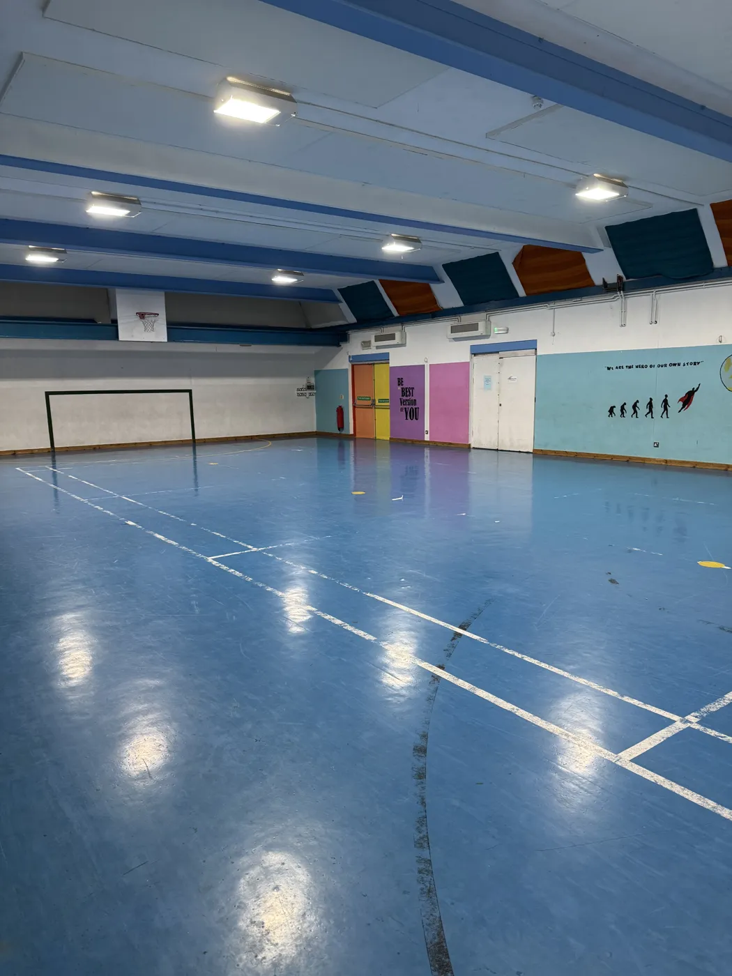 The sports hall at Roots4Learning, with a blue floor, court markings, a goal and a painted mural on the far wall