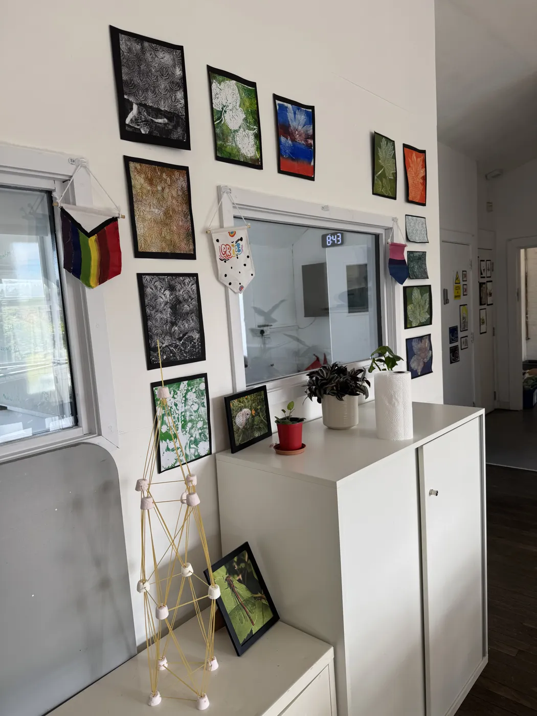 Framed student artwork and photographs hung on a white wall above a cupboard with plants at Roots4Learning