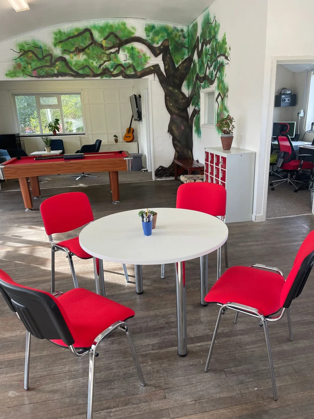 The main room at Roots4Learning: red chairs round a white table, a pool table, a guitar and a painted tree mural across the wall