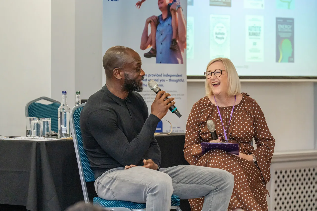 Francis Duku speaking into a microphone while Sue Doughty laughs beside him on stage at the Westmeria Mental Health Summit