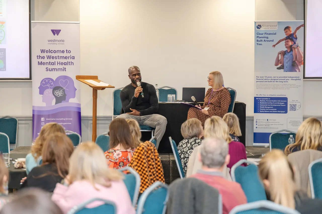 Audience seated in front of the stage as Francis Duku and Sue Doughty talk, with a Westmeria Mental Health Summit banner on the left