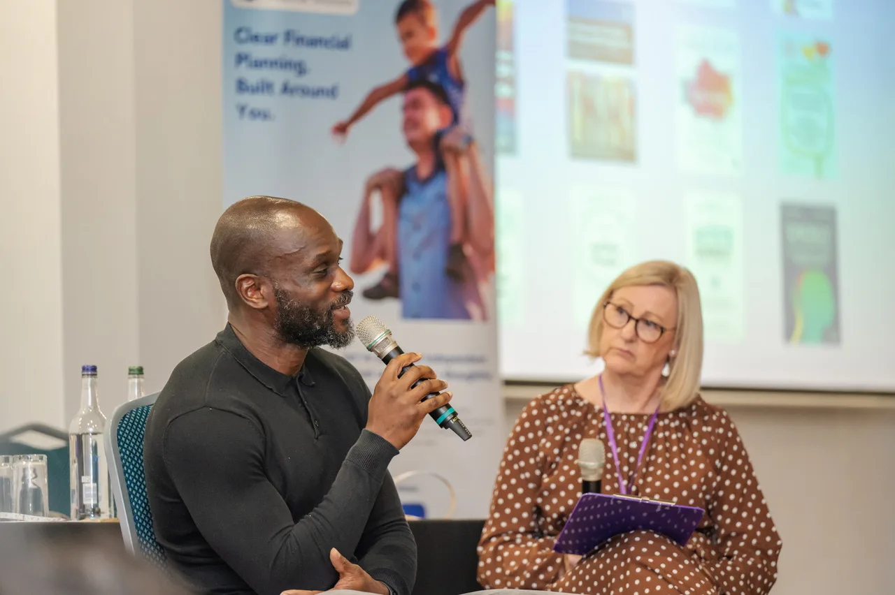 Francis Duku holding a microphone as Sue Doughty listens, holding a purple folder